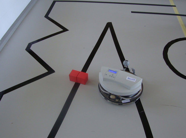 Используя датчики света, робот движется строго по черной линии-зигзагу. Если на пути появляется препятствие, он должен сам, пользуясь только заданной программой, обойти красные кубики и вернуться на черную дорожку